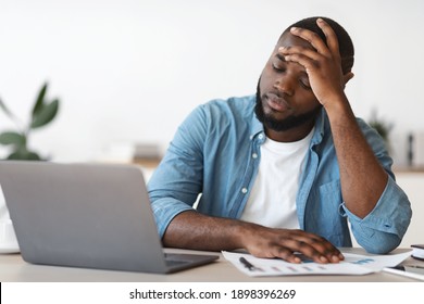 Stressed Black Male Entrepreneur Having Problems At Work. Depressed African American Man Sitting Upset At Desk With Laptop In Office And Touching Head In Despair, Closeup Shot With Free Space