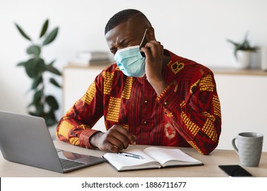 Stressed Black Man In Protective Medical Mask Scratching Face, Having Itchy Skin, Suffering From Allergy At Workplace, African American Guy In Traditional Shirt Sitting At Desk With Laptop In Office