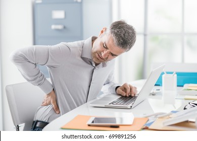 Stressed businessman with backache, he is working at office desk and massaging his back