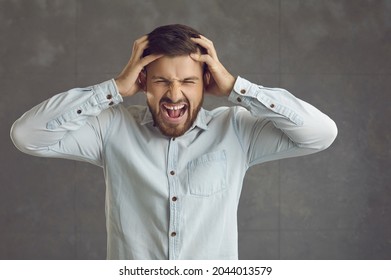 Stressed Businessman Going Postal. Mad Guy Shouting And Tearing Hair Out. Portrait Of Angry Young Man Screaming And Pulling Hair Out Standing On Gray Studio Background. Stress, Rage And Anger Concept