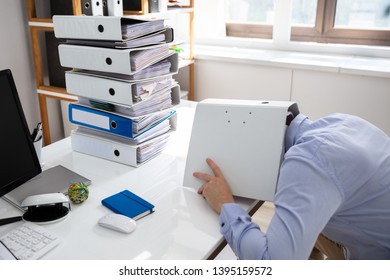 Stressed Businessman Hiding Under Folder While Working At Desk