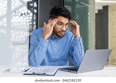 A stressed businessman holding his head while working on a laptop in a modern office. Concept of stress, workload, and business challenges. - Powered by Shutterstock