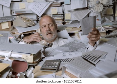 Stressed businessman overwhelmed by work, he is drowning under a lot of paperwork and answering phone calls - Shutterstock ID 1811589811
