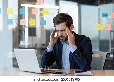 Stressed businessman sitting at office desk working on laptop, holding his head, experiencing a headache or frustration. Office environment with sticky notes and documents. Concept of work stress and - Powered by Shutterstock