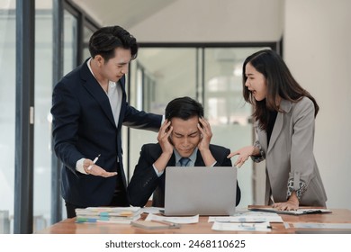 Stressed businessman surrounded by colleagues arguing while working on a laptop in a modern office setting, highlighting workplace tension. - Powered by Shutterstock