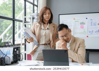 Stressed businessman working with documents and digital tablet at the office with a female colleague bringing more work - Powered by Shutterstock