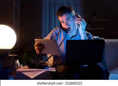 Stressed Businessman Working Overtime Late At Night In The Living Room With Telephone, Computer And Tablet.