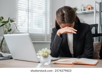 A stressed businesswoman in a suit sits at a desk with a laptop and open book, covering her face in frustration.