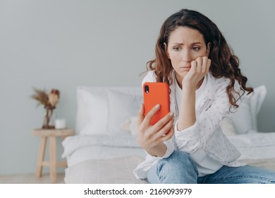 Stressed Caucasian Girl Is Reading Morning News Looking At Smartphone Display. Young Woman Is Chatting On Phone Or Browsing Internet At Home In Her Bedroom. Lady Is Doom Scrolling.