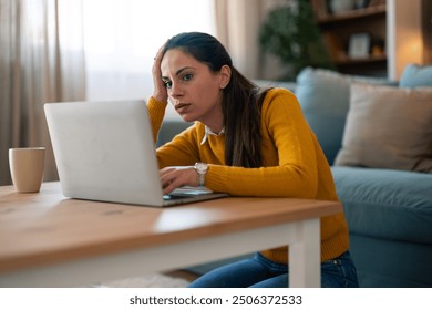 A stressed Caucasian woman in casual attire is focused on her laptop, seated on the floor by a coffee table in a living room, signaling a challenging moment during remote work. - Powered by Shutterstock