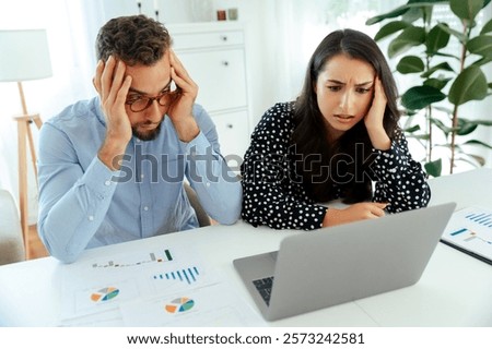 Stressed colleagues analyzing data and charts on a laptop and printed sheets at a desk, working together to solve a problem while sitting in a modern office, experience confusion and disappointment