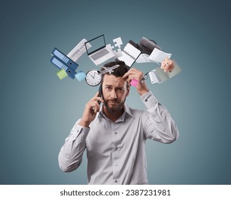 Stressed confused businessman having a phone call, he is surrounded by business items and office supplies: efficiency, planning and stress management concept