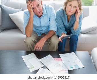 Stressed Couple Sitting On Their Couch Paying Their Bills Smiling At Camera At Home In The Living Room