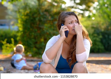 Stressed Depressed Young Mother Talking On A Mobile Phone As She Sits Outside In A Playground With Her Baby Son Playing Behind Her