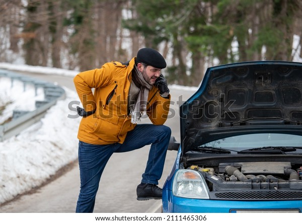 Stressed driver stand on\
the road next to his broken car and looks at the engine on cold\
weather with snow