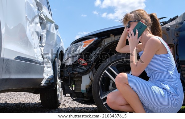 Stressed driver talking on\
sellphone on roadside near her smashed vehicle calling for\
emergency service help after car accident. Road safety and\
insurance concept