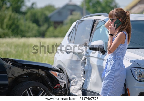 Stressed driver talking on\
sellphone on roadside near her smashed vehicle calling for\
emergency service help after car accident. Road safety and\
insurance concept