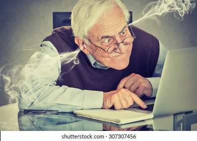 Stressed elderly old man using computer blowing steam from ears. Frustrated guy sitting at table working on laptop isolated on gray wall background. Senior people and technology concept  - Powered by Shutterstock