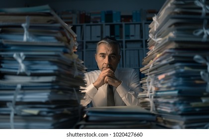 Stressed Exhausted Business Executive Working In The Office Late At Night With Piles Of Paperwork, He Is Overloaded With Work: Management And Deadlines Concept