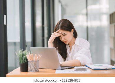 The stressed and exhausted millennial businesswoman is seen sitting at her office desk with her hand on her head, indicating a hard working day where she is overloaded with work - Powered by Shutterstock