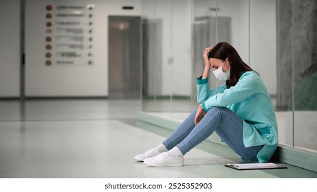 A stressed female healthcare worker in scrubs and a face mask sits on the floor, head in hand, appearing overwhelmed. A clipboard lies beside her in a sterile hospital hallway, symbolizing burnout. - Powered by Shutterstock