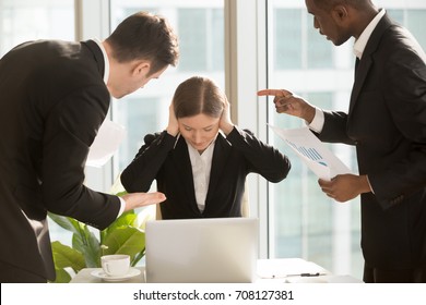 Stressed Female Office Worker Closing Ears With Hands While Sitting At Desk Surrounded By Nervous Mad Colleagues Or Bosses Pointing At Mistakes In Report, Demanding Do Job Faster. Criticism At Work 