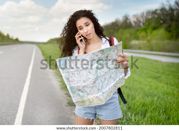Stressed female tourist with map standing on\
roadside, speaking on smartphone, cannot catch car or find her\
travel route. Young lady feeling lost while hitchhiking on highway,\
having problem