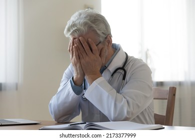 Stressed frustrated mature doctor wearing white uniform with stethoscope covering face with hands, sitting at work desk in hospital, sad senior gp feeling desperate and guilty for medical failure - Powered by Shutterstock