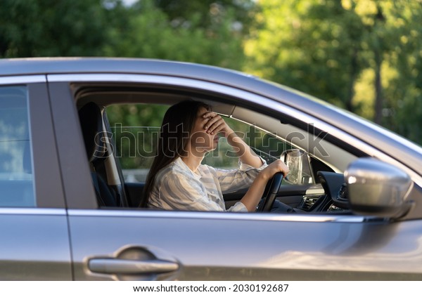 Stressed girl with headache driving car touching
forehead with hand. Frustrated young female driver suffering from
illness or hangover, displeased with heat inside vehicle, tired
from stress overwork
