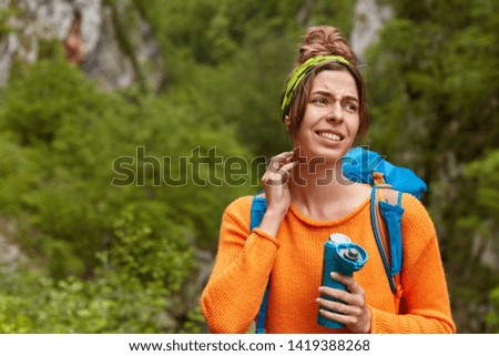 Stressed girl traveler get lost in forest, cannot find way, looks with upset expression aside, poses on green natural background, drinks tea from flask, wears rucksack, orange jumper, feels worried