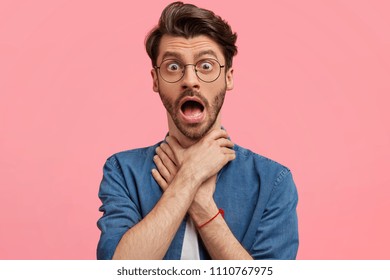Stressed good looking male suffers from asthma or suffocation, keeps hands on neck and opened mouth, wears round spectacles, dressed in denim shirt, stands against pink background, has obstruction