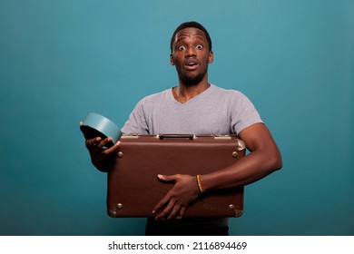 Stressed Guy Carrying Suitcase And Running Late For Vacation Trip, Looking At Time On Clock Over Blue Background. Person With Retro Briefcase Having Watch To Prepare For Work Voyage.