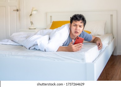 Stressed handsome Asian man sleeping on the bed awakening by smartphone alarm clock ringing and shocked. Tired guy office worker wake up late in the morning.