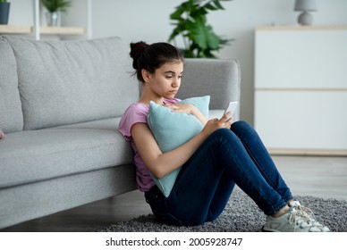 Stressed Indian Teen Girl Sitting On Floor With Cellphone And Reading Bad Message At Home. Upset Adolescent With Mobile Phone Feeling Sad, Suffering From Cyber Bullying, Being Harassed Online