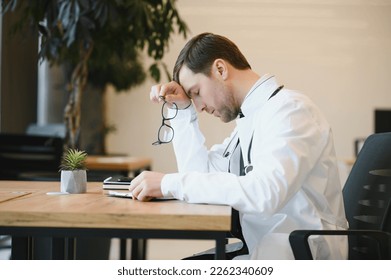 Stressed male doctor sat at his desk. Mid adult male doctor working long hours. Overworked doctor in his office. Not even doctors are exempt from burnout - Powered by Shutterstock