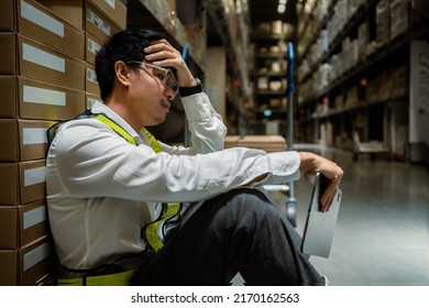 A Stressed Male Manager Or Worker Sits Leaning Against A Box Holding A Tablet And In A Warehouse. Concept Of Thinking About Problems At Work. Supply Chain And Warehouse Business Concept .