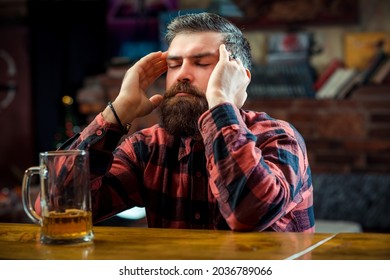 Stressed Man Drinking Beer At Pub. Alone Bearded Man Sitting At The Bar Counter. Alcohol Addiction. Bearded Man Holds Glass Of Beer. Tired Brutal Guy With Headache In The Beer Pub.