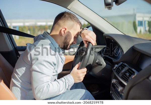Stressed man feeling headache in car, keeping hand to\
head and feeling anxiety. Driver falling asleep on steering wheel\
of car