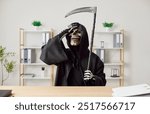 Stressed man in grim reaper death costume with black cloak and scythe sitting at desk and putting hand to forehead. He looking upset at making a mistake or tired from work, gesturing sad expression.