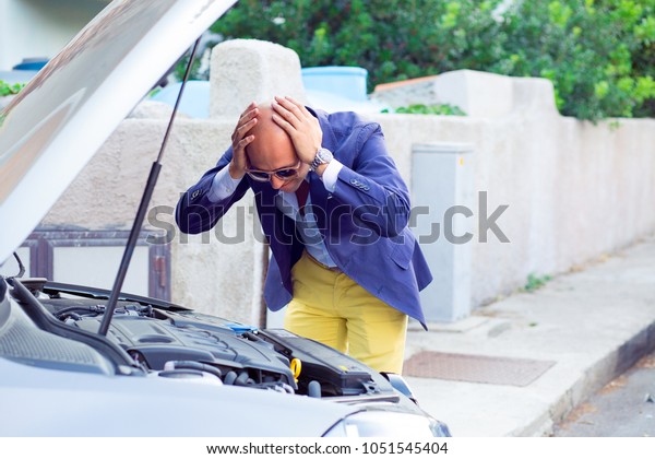 Stressed man having trouble with his broken\
car looking in frustration at failed engine; hands on head looking\
inside to the open capote top of the car. Negativehuman emotion\
reaction body\
language\
