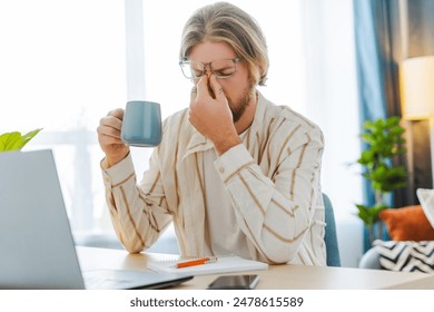 Stressed man in home office with headache and eye strain from remote work, reflecting burnout. Despite health issues, he pushes through, emblematic of challenges faced by digital nomads