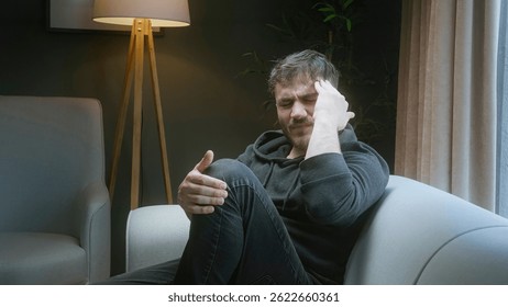 Stressed Man Sitting on Couch Holding His Head and Knee in Pain at Home