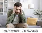 Stressed man sitting on sofa holding his head, feeling overwhelmed and anxious. Man experiencing mental health issues, worry, frustration, and emotional stress. Concept of anxiety and mental health