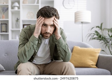 Stressed man sitting on sofa holding his head, feeling overwhelmed and anxious. Man experiencing mental health issues, worry, frustration, and emotional stress. Concept of anxiety and mental health