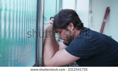 Stressed man struggling with mental illness leaning by window suffering from emotional despair. One worried male person in 30s feeling pressure