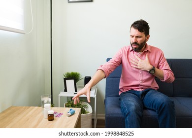 Stressed Man Suffering An Asthma Attack And Having Trouble Breathing. Young Man Reaching For His Inhaler While Sitting On The Sofa