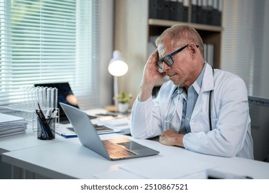 Stressed mature male doctor in white coat sitting at desk in office looking concerned at laptop screen - Powered by Shutterstock