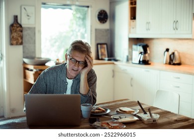 Stressed mature man using laptop on kitchen table during breakfast - Powered by Shutterstock