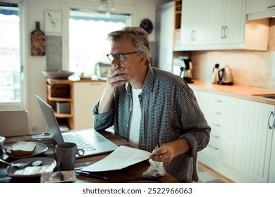 Stressed mature man using laptop on kitchen table during breakfast - Powered by Shutterstock