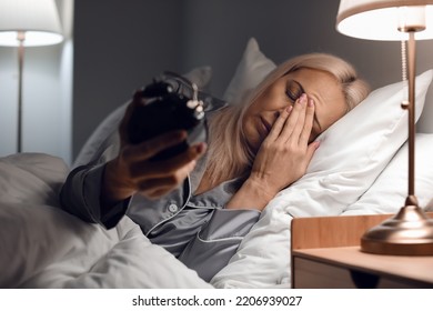 Stressed mature woman with alarm clock lying in bed at night - Powered by Shutterstock